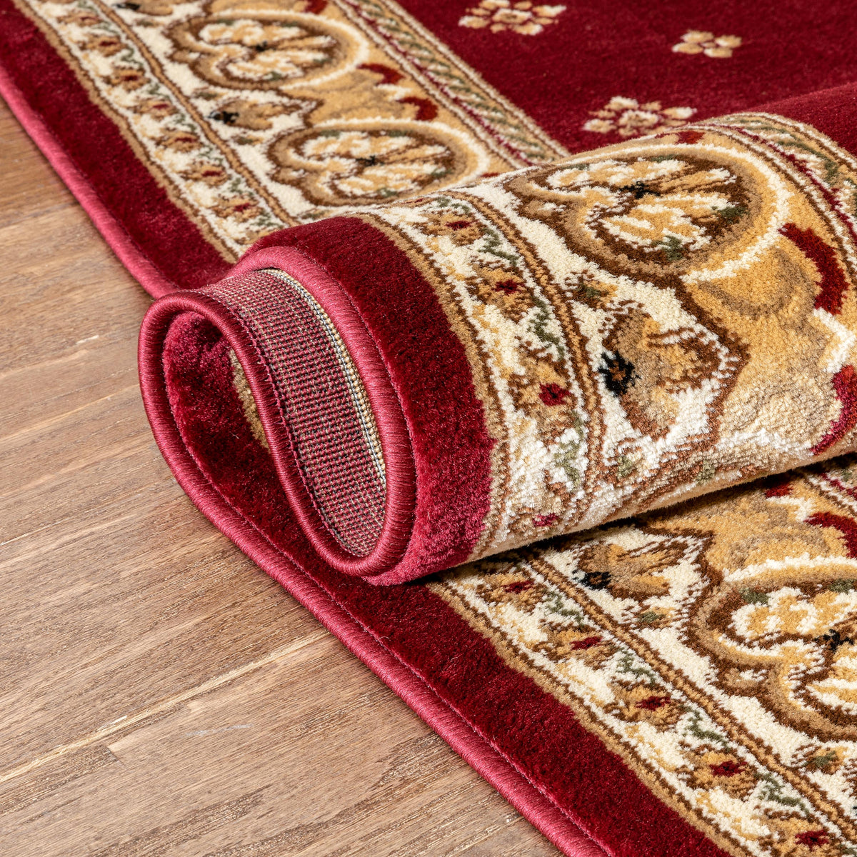 A close-up of a rolled area rug with a traditional ornate pattern in deep red, beige, and gold, shown on a light brown wood floor.
