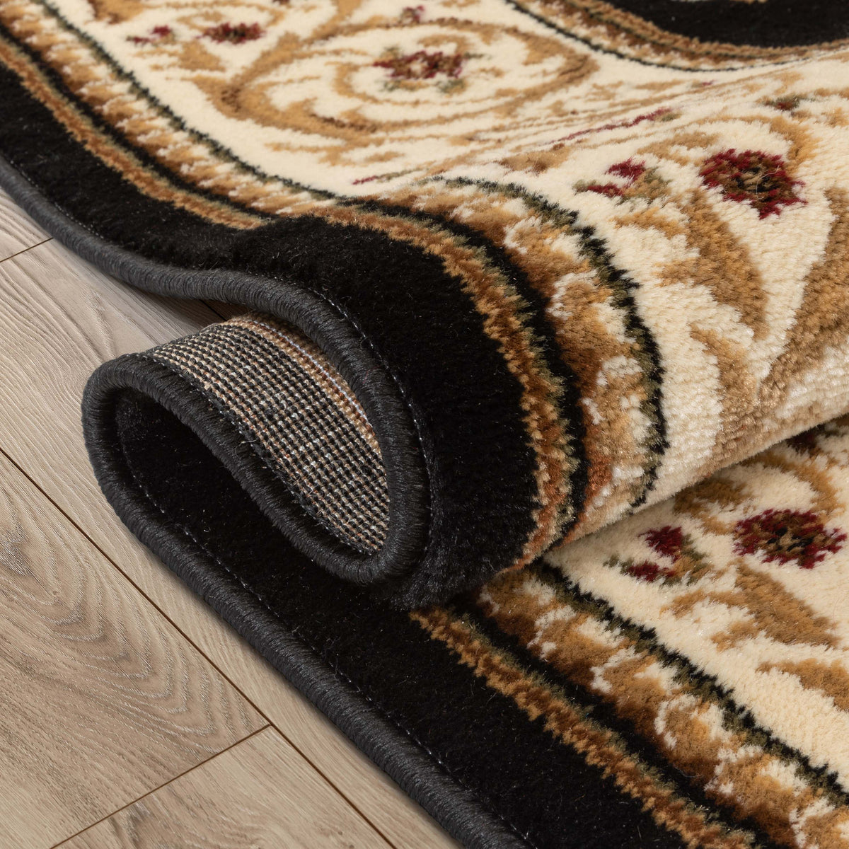 A close-up of a rolled area rug showing its cream and tan patterned field, dark brown floral accents, and a thick black border, placed on a light wood floor.