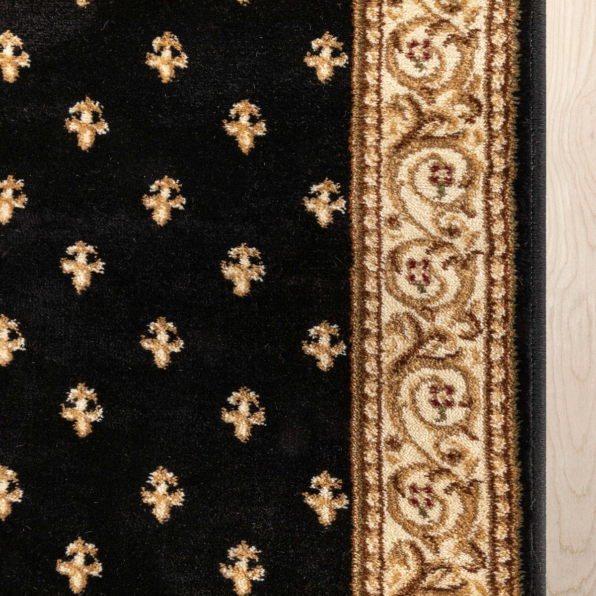 Close-up of a black rug with repeating gold medallion motifs and a wide ornate gold border, shown against a light wood floor.
