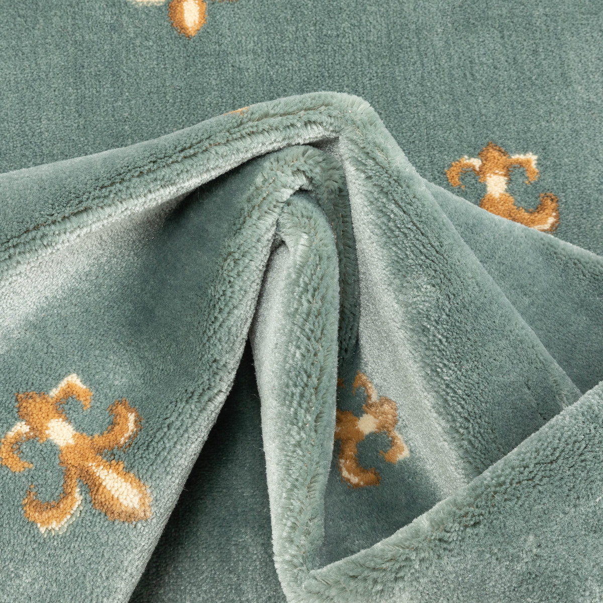 A close-up of a plush, dusty sage green fabric, likely a throw blanket or fleece, featuring embroidered golden-brown fleur-de-lis motifs.