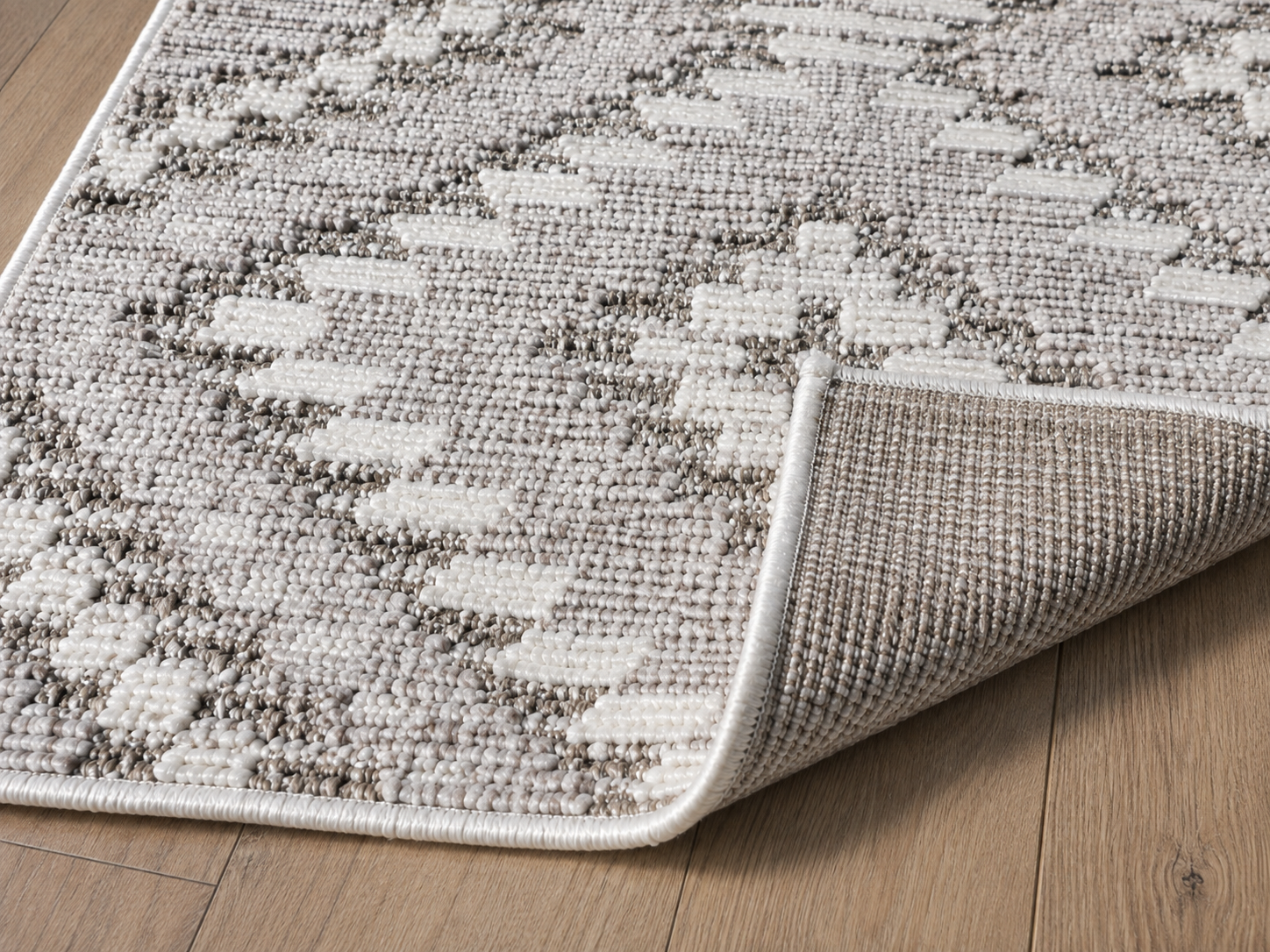 A close-up of a textured area rug with a beige and off-white geometric pattern, shown with one corner folded over to reveal the backing, placed on a light brown wood floor.