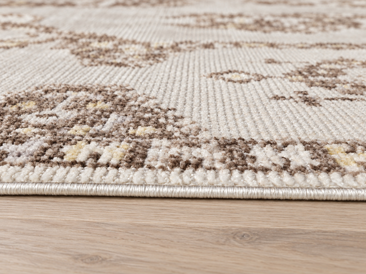 Close-up of a textured area rug with a beige and brown traditional pattern, shown on a light wood floor.