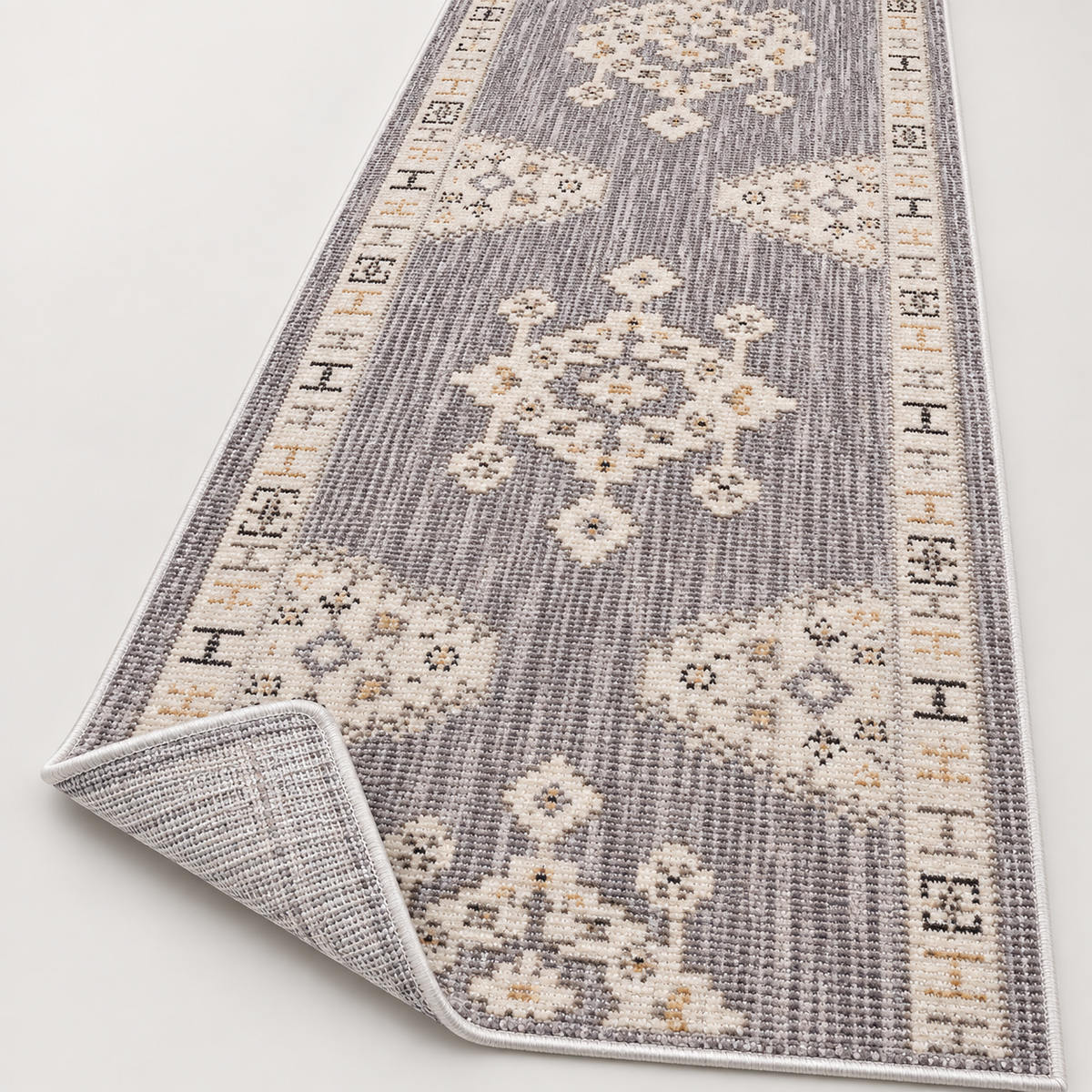 A long rectangular area rug shown on a light neutral background, with one corner folded back to reveal the underside. The rug has a gray and off-white woven pattern with cream and tan medallion motifs and decorative borders.