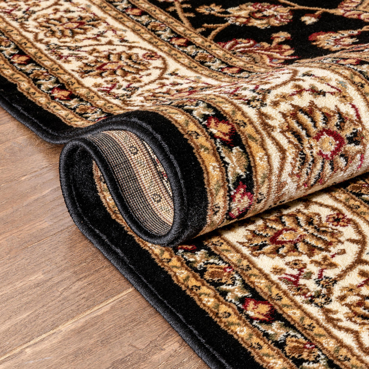 A close-up of a rolled area rug with an intricate traditional floral pattern in beige, black, brown, and muted red, shown on a wood floor.
