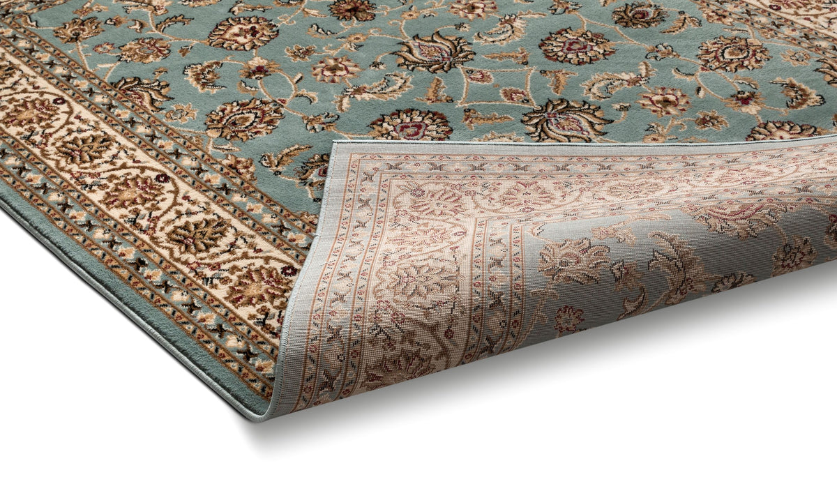 A traditional oriental-style area rug with a dusty teal-blue field, beige and brown floral motifs, and a matching patterned border, shown with one corner rolled up on a white background.