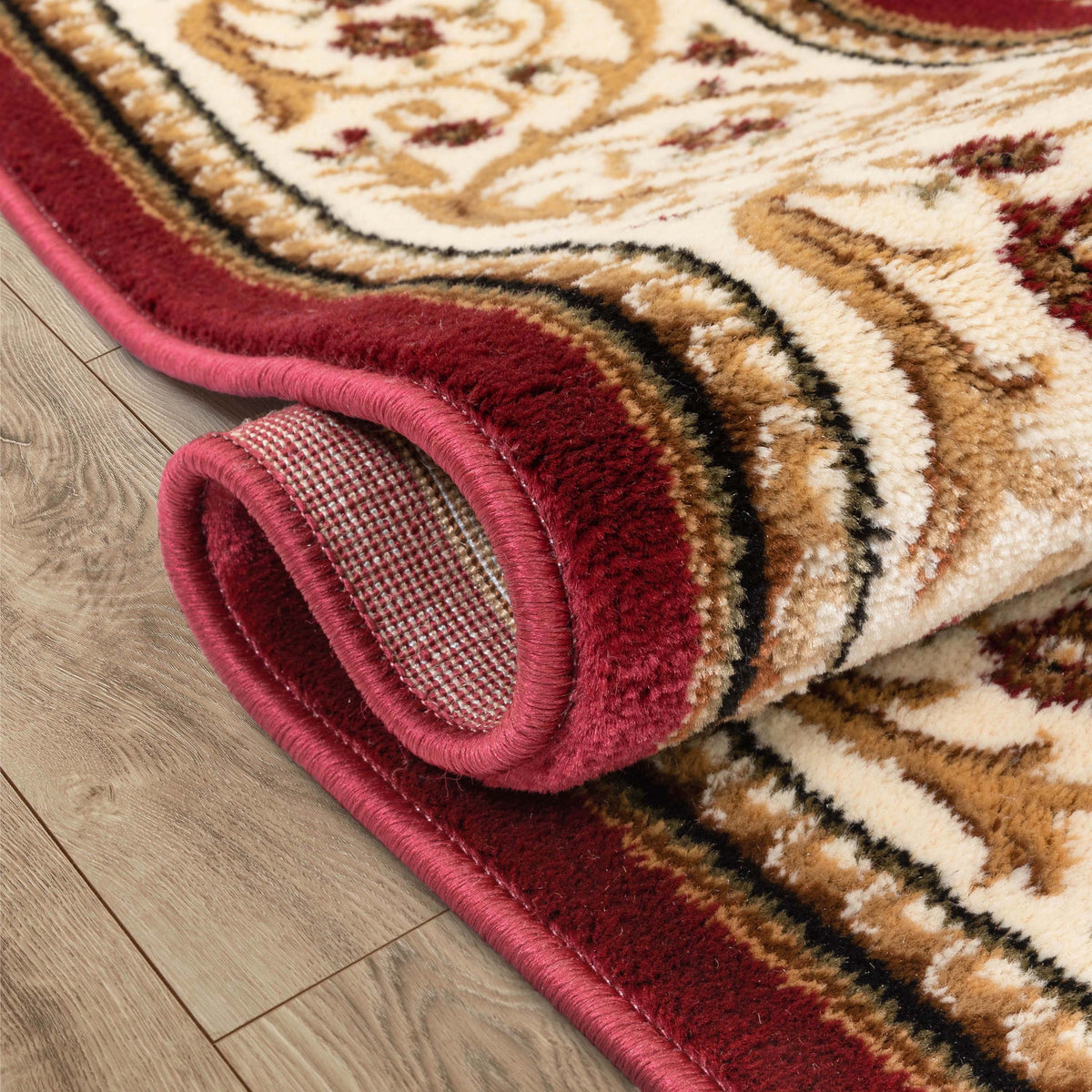 A close-up of a rolled area rug with a traditional pattern, featuring a deep red border and cream, tan, and burgundy motifs, shown on a light wood floor.