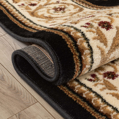 A close-up of a rolled area rug showing its cream and tan patterned field, dark brown floral accents, and a thick black border, placed on a light wood floor.