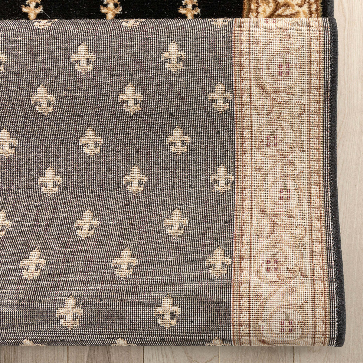 A close-up of a woven rug or textile showing a dark gray field with repeating cream fleur-de-lis motifs and a wide cream border with ornate floral patterns and small rust accents, laid against a light wood floor.