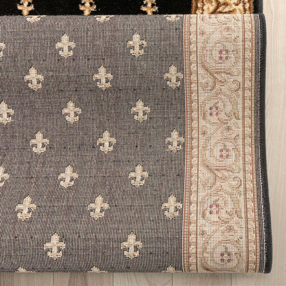 A close-up of a woven rug or textile showing a dark gray field with repeating cream fleur-de-lis motifs and a wide cream border with ornate floral patterns and small rust accents, laid against a light wood floor.