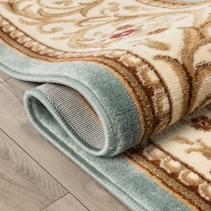 A rolled section of a traditional area rug with a beige and brown floral scroll pattern and a dusty blue-green border, shown on a light gray wood plank floor.