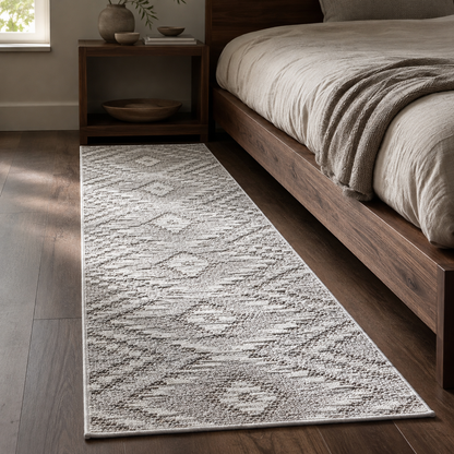 A long runner rug with a textured geometric diamond pattern in off-white and light gray, styled in a bedroom next to a wooden bed frame and nightstand.
