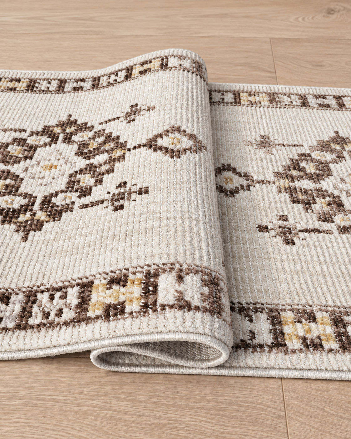 A close-up of a folded area rug with a textured woven pattern in cream, brown, and tan, displayed on a light wood floor.
