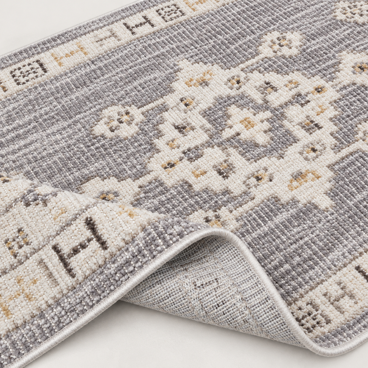 A close-up of a woven area rug with a gray and cream geometric medallion pattern, showing the underside and folded corner on a light neutral background.