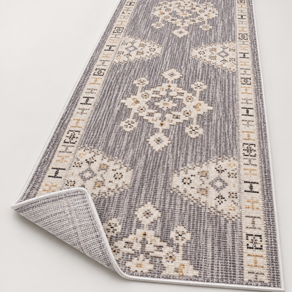 A long rectangular area rug shown on a light neutral background, with one corner folded back to reveal the underside. The rug has a gray and off-white woven pattern with cream and tan medallion motifs and decorative borders.