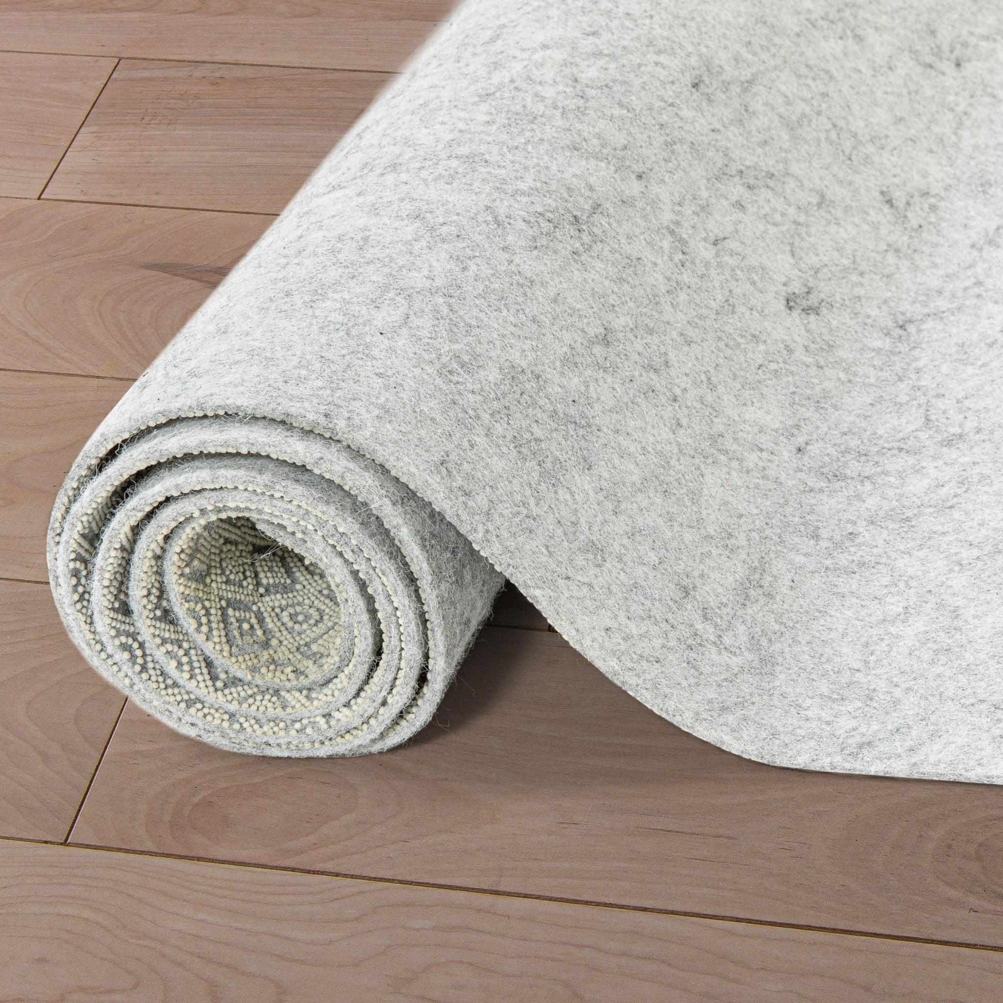 PAD-1 - Well Woven Premium Felt Non-Slip Rug Pad - Rolled-up gray rug pad 1/4" thick on a wooden floor. The top of the pad is felt and the bottom is non-slip rubber